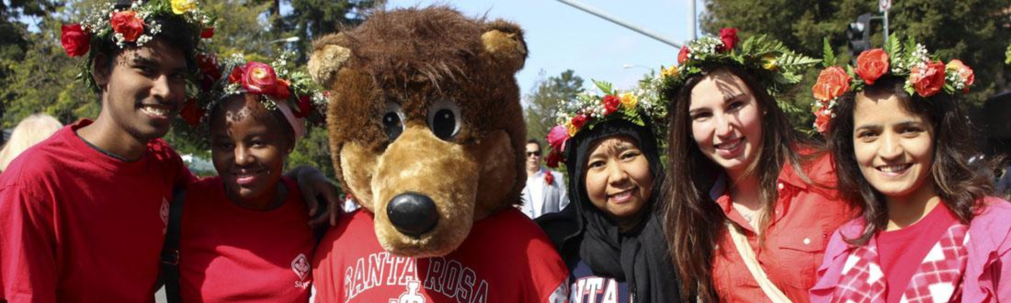 Decorative banner with five students surrounding Rosco on a sunny day, wearing red shirts and flower hats